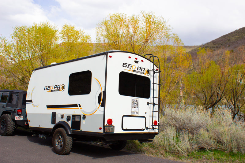 A white Geo Pro RV rental trailer hitched to a dark grey Jeep Wrangler, parked roadside amidst vibrant spring foliage. — photo 1