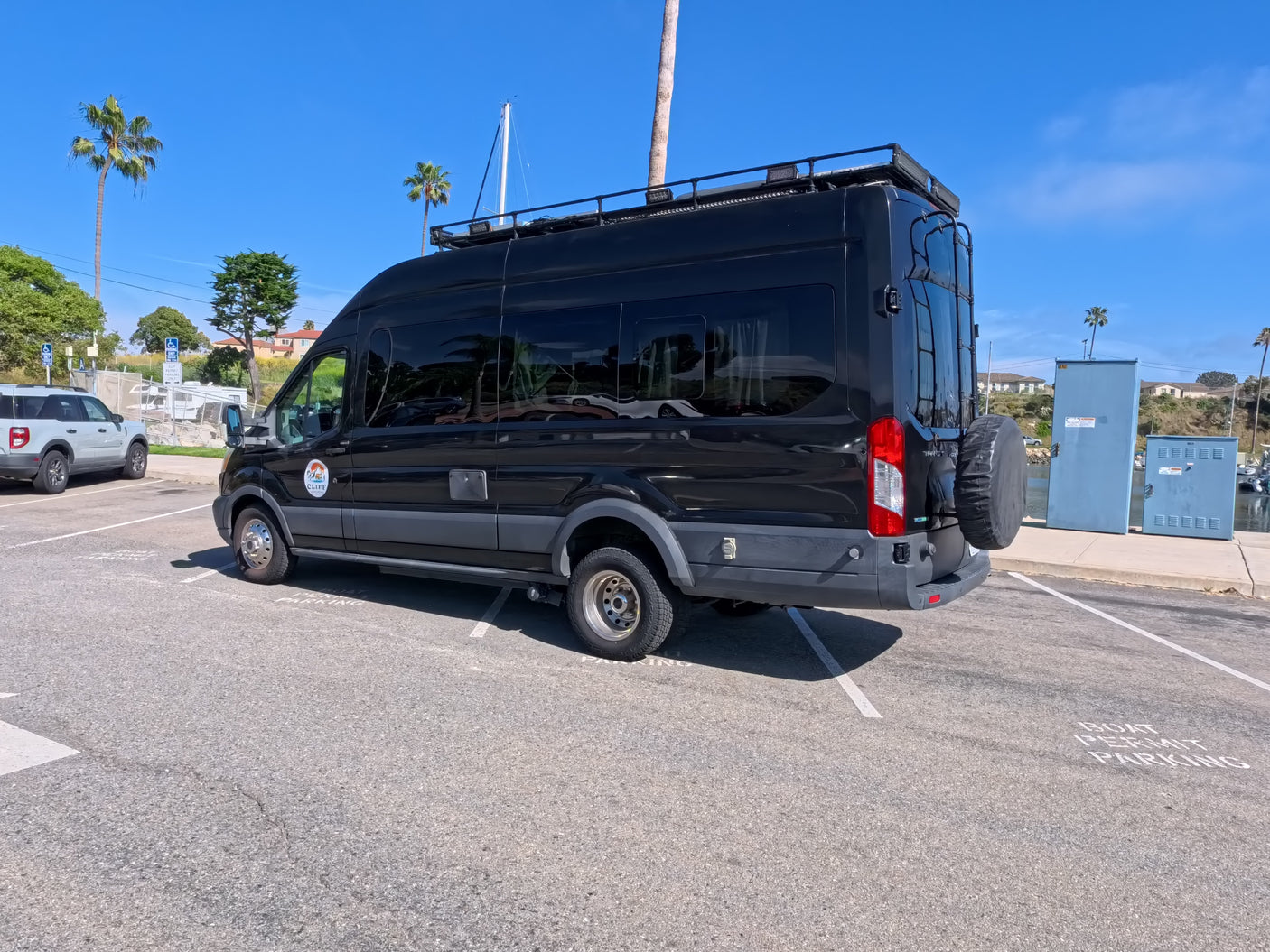 Black rv rental camper van with adventure gear, parked by a sunny marina in 'Boat Permit Parking'.