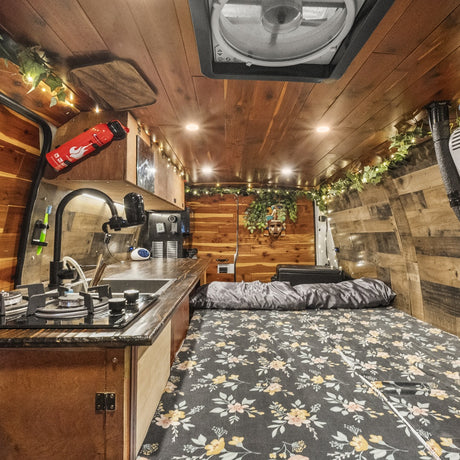 This RV rental shows a cozy, wood-paneled camper van interior with a kitchen (sink, stove) and a floral-patterned bed.