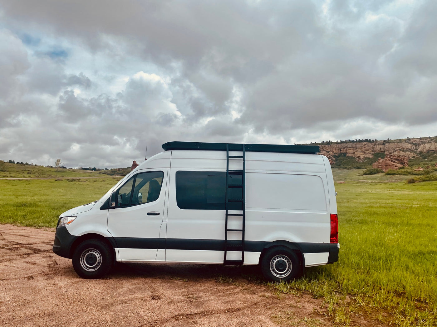 White Sprinter camper van with roof rack & ladder, parked on dirt next to green fields and red rocks. Ideal RV rental. — photo 10