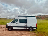 White Sprinter camper van with roof rack & ladder, parked on dirt next to green fields and red rocks. Ideal RV rental.