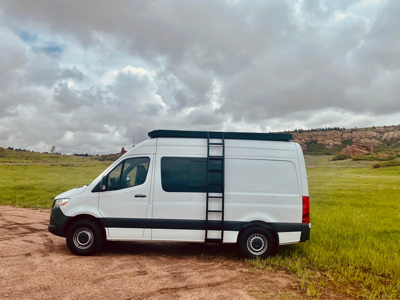 White Sprinter camper van with roof rack & ladder, parked on dirt next to green fields and red rocks. Ideal RV rental. — photo 10