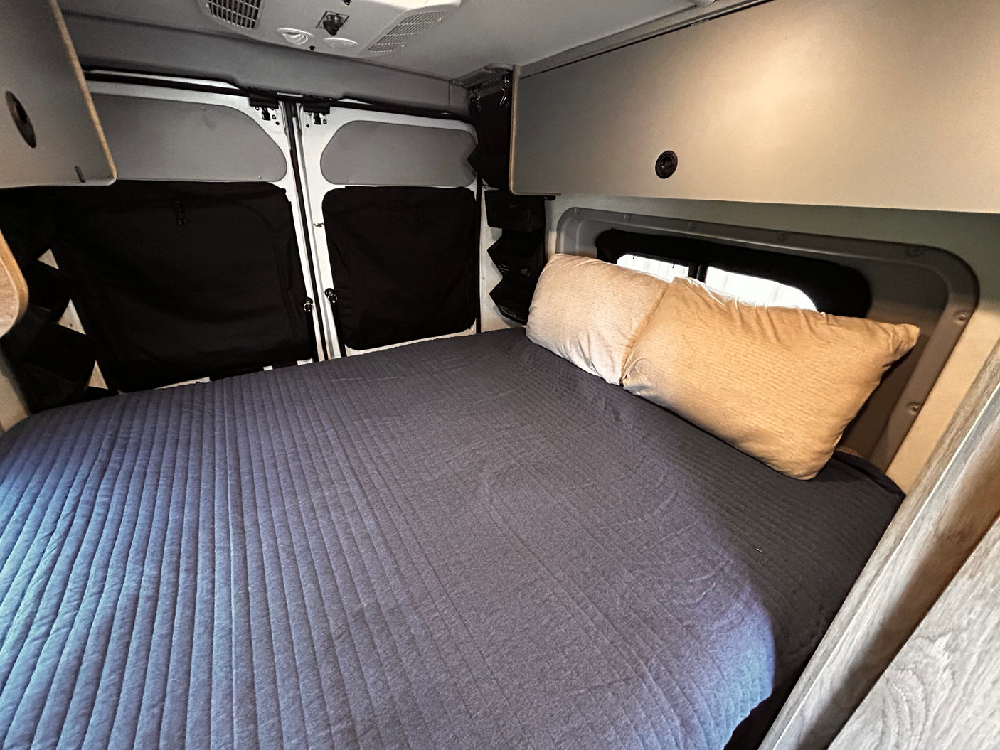 This RV rental's interior features a cozy bed with pillows, window coverings, and overhead storage.
