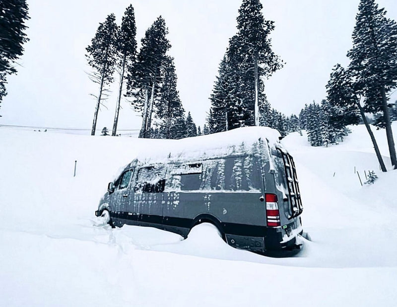 An RV rental van is deep in snow, almost buried, amidst pine trees in a vast winter landscape. — photo 11