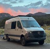This beige Mercedes Sprinter RV rental is set up for adventure, parked at sunset against scenic mountains with a campfire nearby.