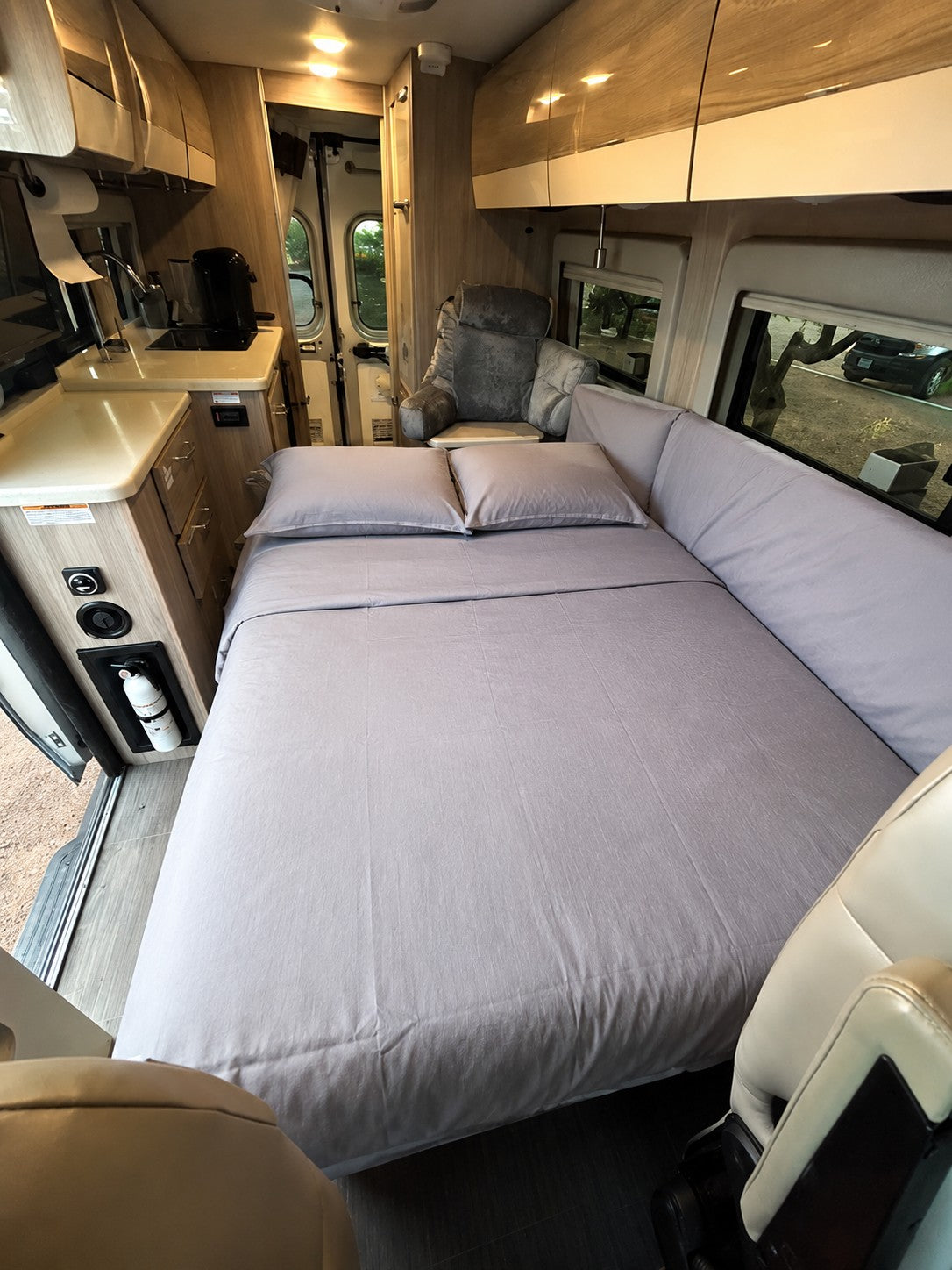Cozy RV rental interior featuring a comfortable bed with gray linens, a compact kitchen, and an armchair for relaxation.