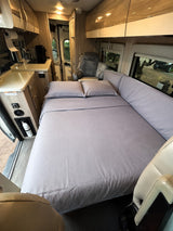 Cozy RV rental interior featuring a comfortable bed with gray linens, a compact kitchen, and an armchair for relaxation.
