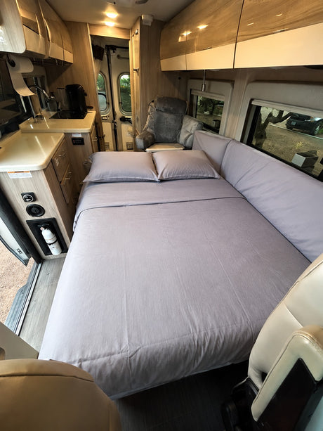 Cozy RV rental interior featuring a comfortable bed with gray linens, a compact kitchen, and an armchair for relaxation.