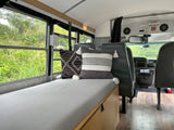 Experience our RV rental: a spacious bus camper with a cozy striped bed, decorative pillows, and nature views.