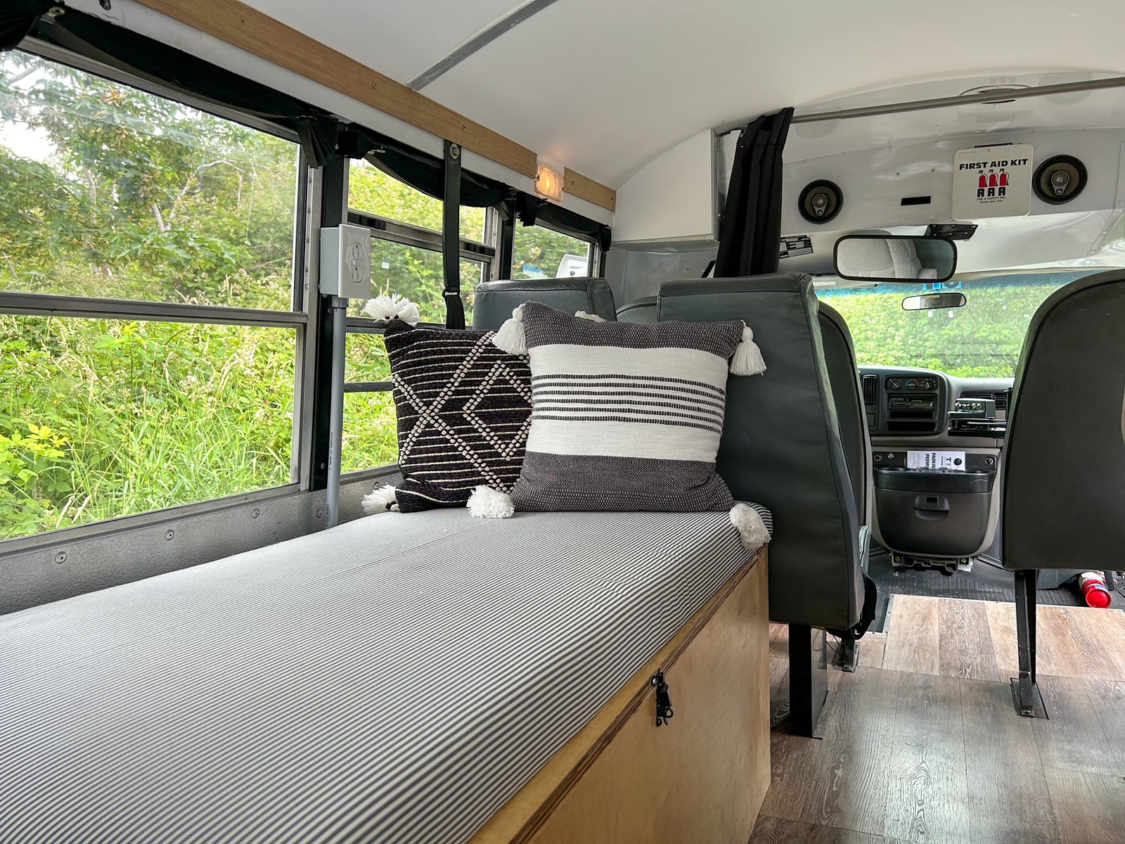 Experience our RV rental: a spacious bus camper with a cozy striped bed, decorative pillows, and nature views.