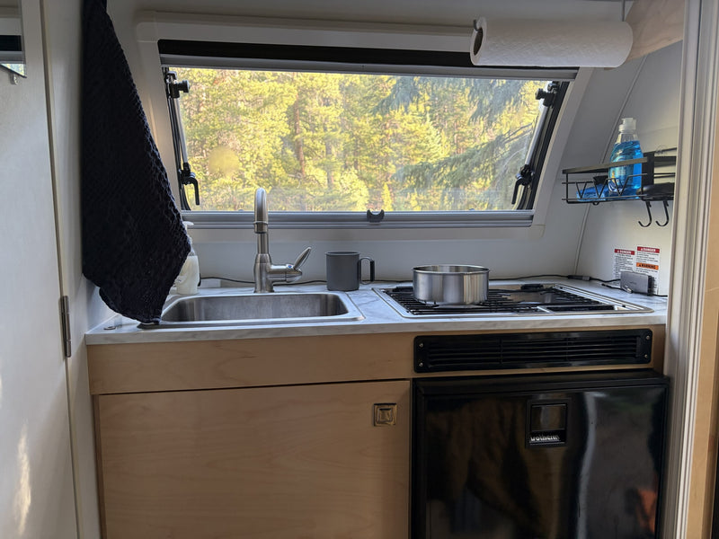  with a dog peeking from the car window.|nuCamp T@B 320 Boondock travel trailer RV rental with extended awning & outdoor kitchen in a forest campsite.|Compact RV rental kitchen: sink — photo 4