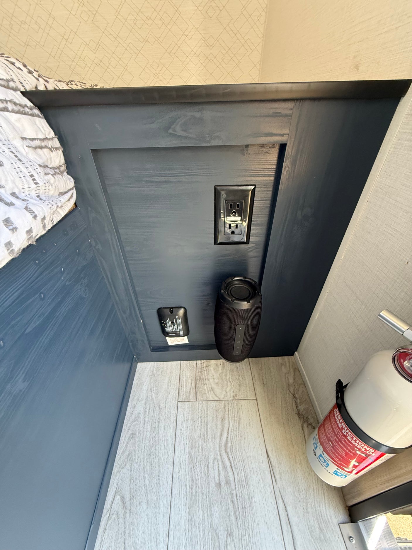 Inside an RV rental bedroom nook: includes a GFCI outlet, CO detector, speaker, and fire extinguisher by light wood flooring.