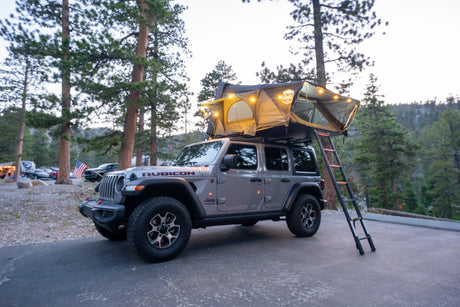 Adventure awaits! A gray Jeep Wrangler with a cozy, lit rooftop tent setup at a beautiful RV rental campsite.