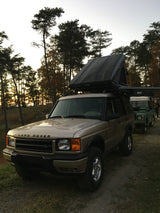 A Land Rover Discovery with an open rooftop tent, perfect for an RV rental adventure, is parked at sunset in the woods. Another Land Rover is seen behind.