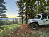 Overland RV rental: White Jeep Rubicon with roof tent on forest hill, overlooking lake at sunset. Perfect adventure.