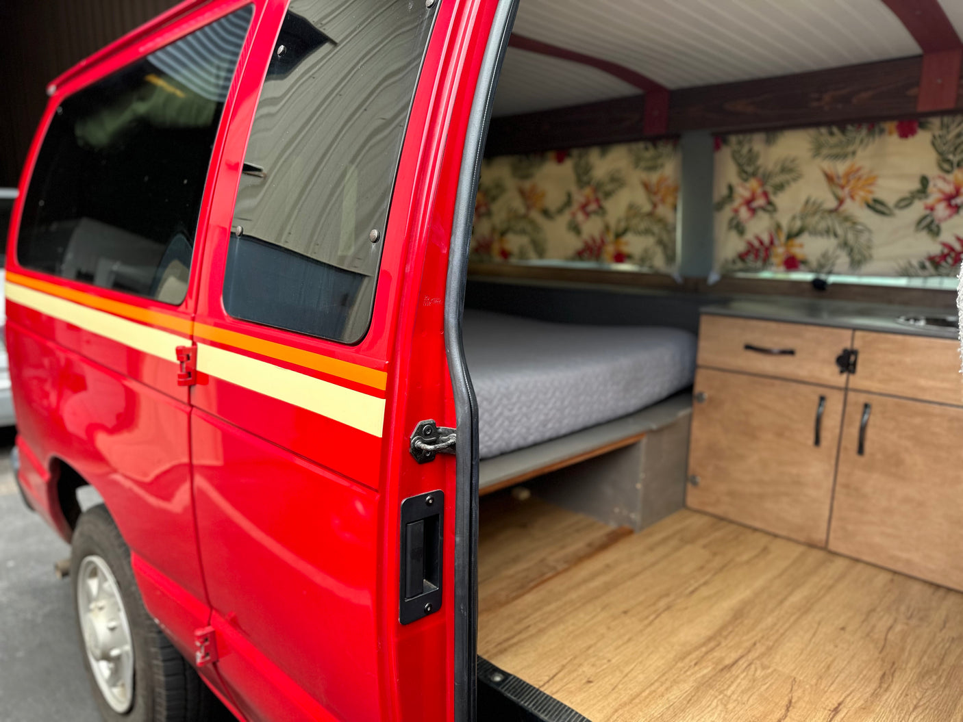 Step inside this custom red camper van, ideal for an RV rental. Features a comfy bed, wood flooring, and kitchen area.