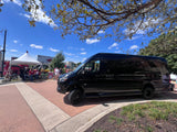 A black Sprinter van, a luxury RV rental, is parked near a stadium and tents on a bright, sunny day with people.