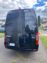 Black Mercedes-Benz Sprinter van, possibly an RV rental, parked on a driveway. Its glossy surface reflects a house and the photographer.