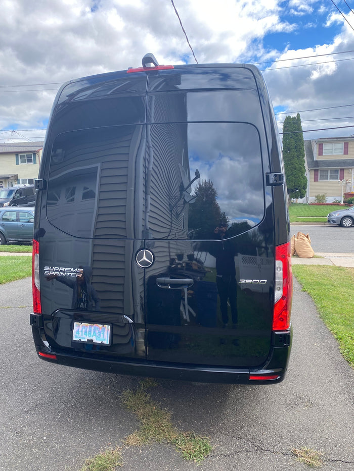 Black Mercedes-Benz Sprinter van, possibly an RV rental, parked on a driveway. Its glossy surface reflects a house and the photographer.
