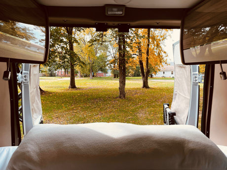View from an rv rental camper van. Pillow in foreground, outside features autumn trees, grassy field, and houses.