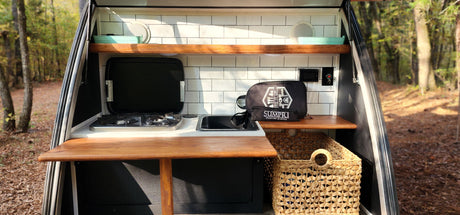 Compact RV rental kitchen features: gas stove, small sink, subway tile backsplash, and wooden shelves. Ideal for outdoor cooking!