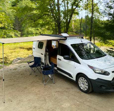 White camper van RV rental with an awning and chairs, perfect for peaceful camping by the forest and pond.