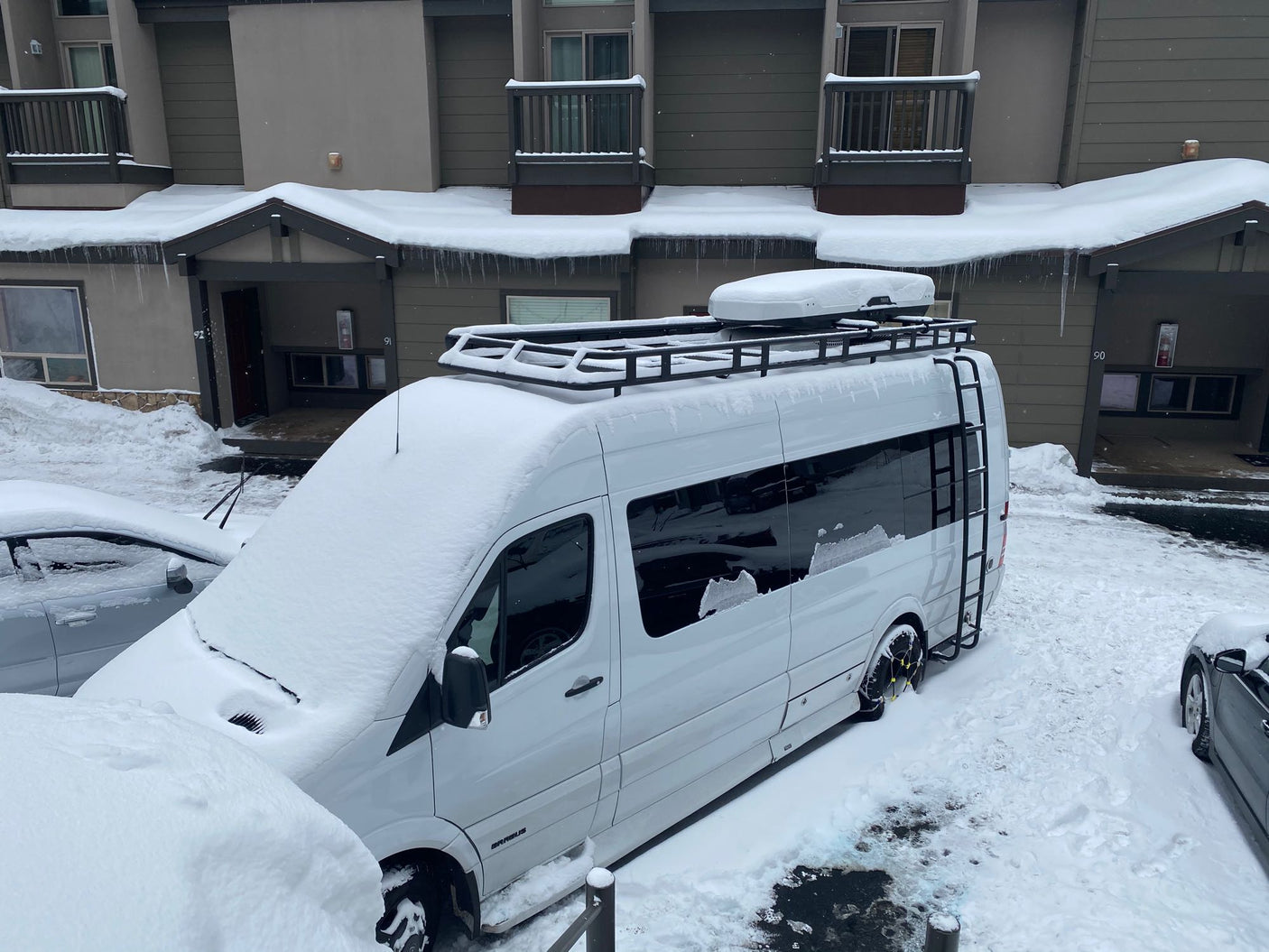 Snow-covered RV rental van with roof rack and chains, parked by a building with icicles after a heavy snowfall.