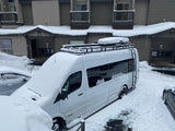 Snow-covered RV rental van with roof rack and chains, parked by a building with icicles after a heavy snowfall.
