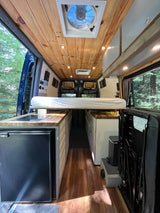 Interior view of a cozy rv rental, showcasing wood paneling, a large vent fan, lighting, and a small kitchen setup with a fridge.