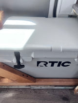 A white RTIC cooler is secured with wooden supports on a dark floor in an RV rental, ready for travel.