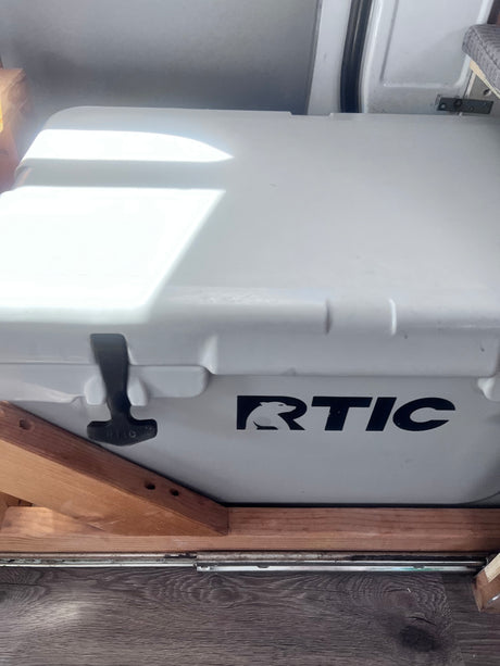 A white RTIC cooler is secured with wooden supports on a dark floor in an RV rental, ready for travel.