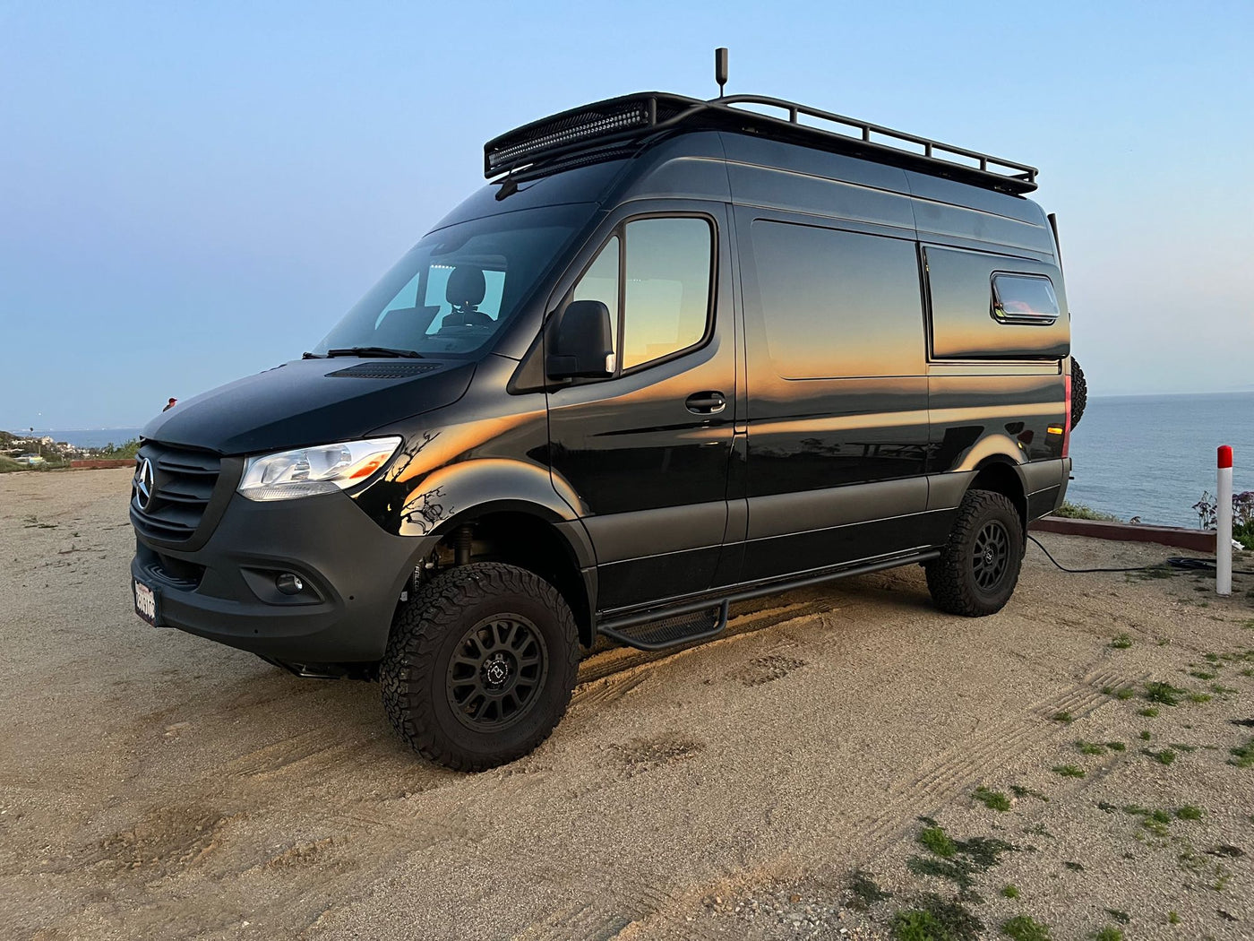 Black Mercedes Sprinter adventure van ready for RV rental, parked on a cliff overlooking the ocean with sunset reflections. — photo 19