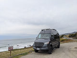Dark gray Mercedes Sprinter RV rental parked at an ocean overlook on a cloudy day, with a scenic coastal road curving uphill.