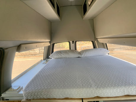 Inside this cozy RV rental camper van: a bed with white pillows, overhead storage, and windows revealing the road outside.