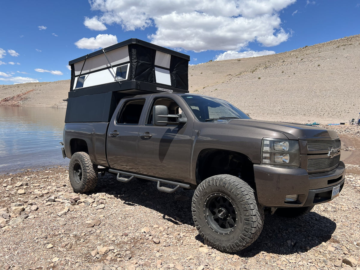 Oru Designs Bruin pop-up truck camper with the roof raised and side windows open, mounted on a lifted Chevrolet Silverado 2500HD parked on a rocky desert lakeshore