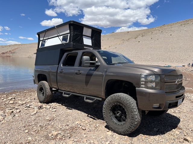 Oru Designs Bruin pop-up truck camper with the roof raised and side windows open, mounted on a lifted Chevrolet Silverado 2500HD parked on a rocky desert lakeshore