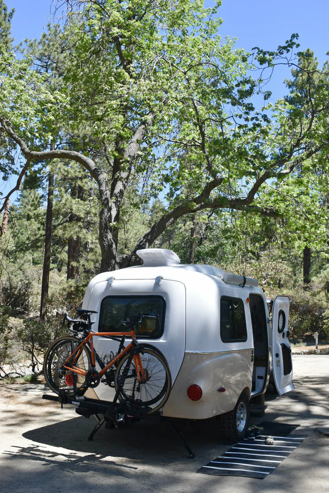 Teardrop RV rental with bikes parked at a sunny forest campsite, ideal for outdoor adventures and exploring nature.