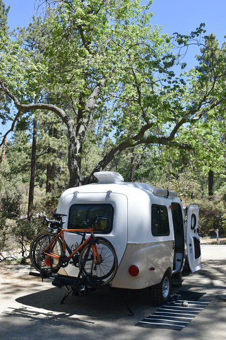 Teardrop RV rental with bikes parked at a sunny forest campsite, ideal for outdoor adventures and exploring nature.