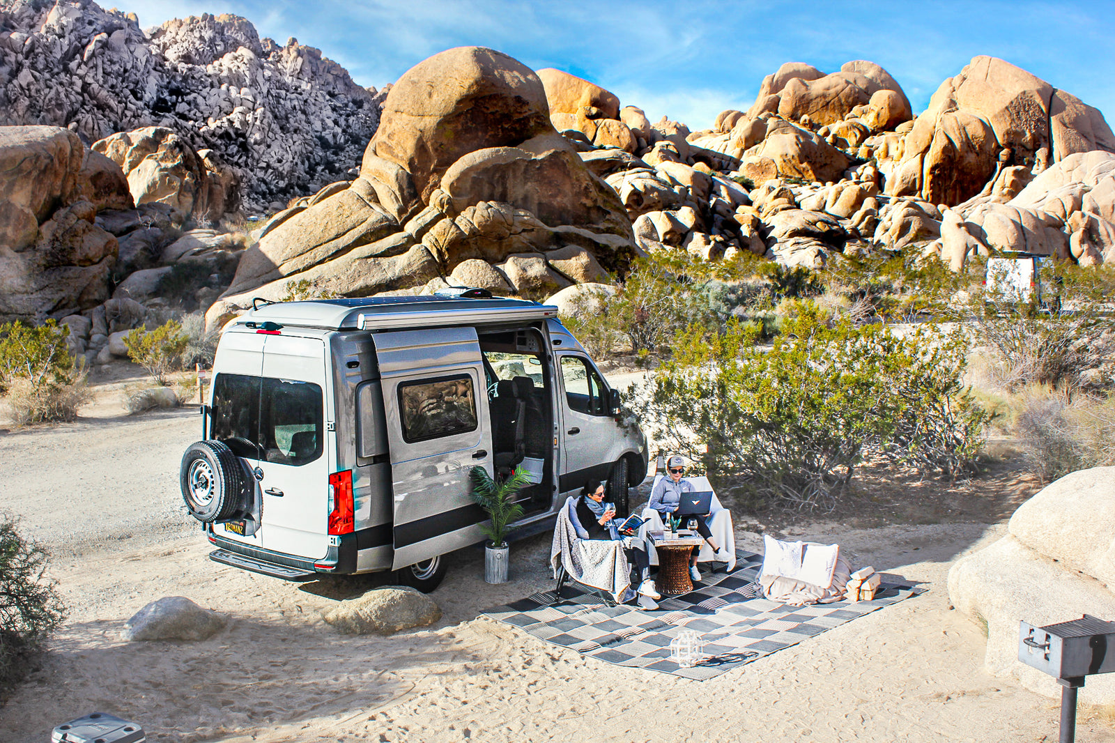 Desert relaxation with an **rv rental**: A camper van, two women reading & computing, surrounded by desert boulders.