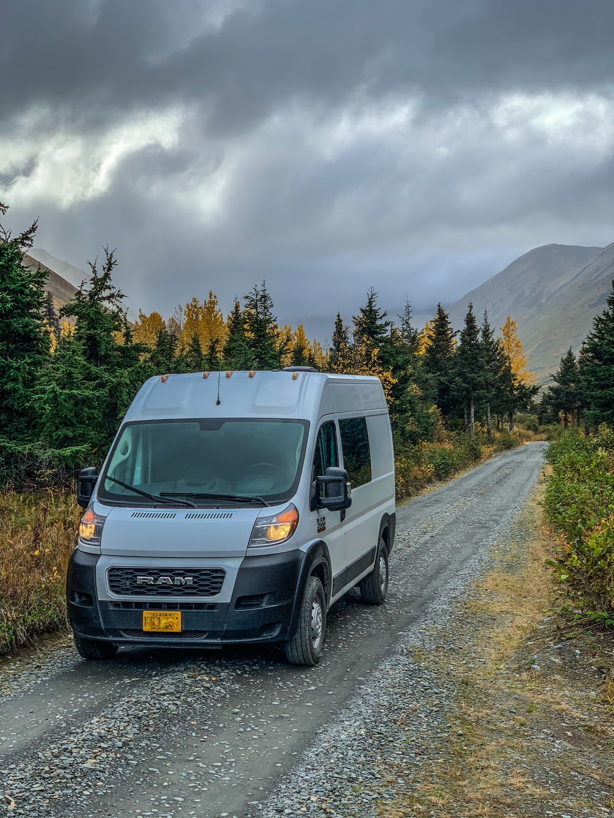 White RV rental van on a gravel road in the Alaskan mountains, surrounded by vibrant autumn trees under a cloudy sky. — photo 9