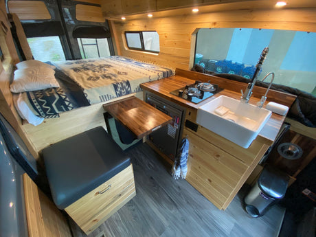 Explore this custom RV rental: comfy bed, compact kitchen with sink/stove, pull-out table, and wood details.