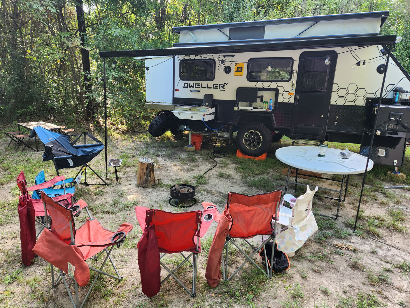 Spacious Dweller 15' RV rental set up at a rustic campsite with chairs, tables, and a fire pit, ready for adventure.