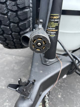 Close-up of a 7-pin RV electrical connector for safe rv rental towing. Part of trailer jack, frame, and rugged tire visible.