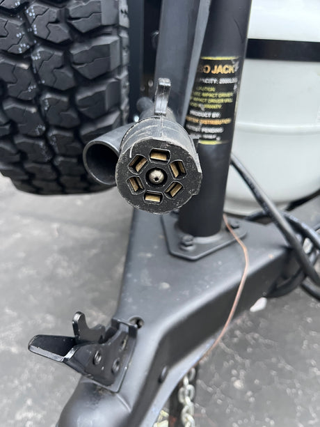 Close-up of a 7-pin RV electrical connector for safe rv rental towing. Part of trailer jack, frame, and rugged tire visible.