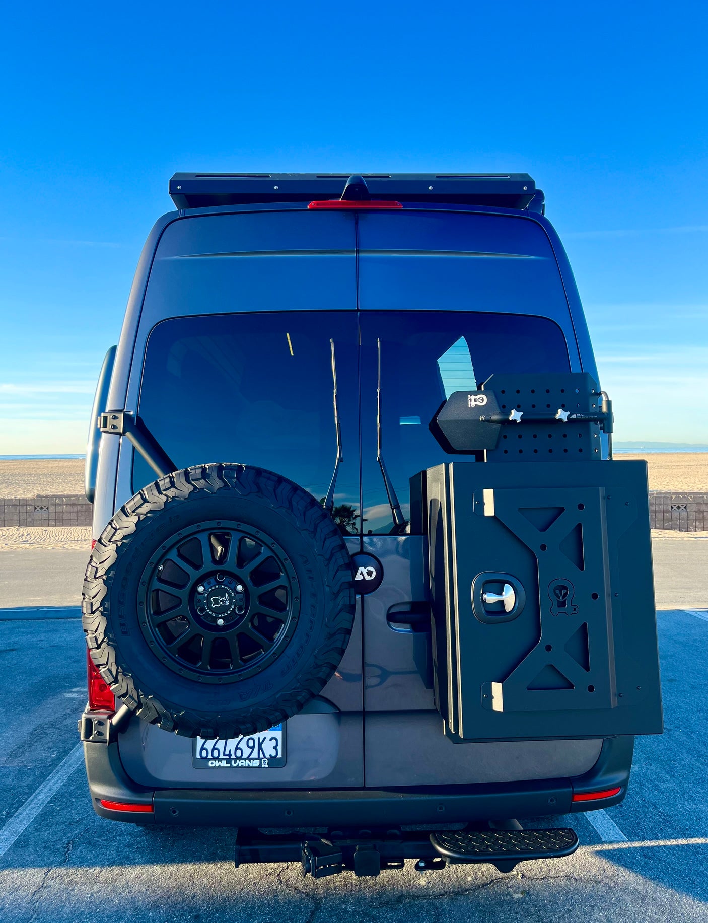 Rear of a gray RV rental van with a spare tire and storage box. Parked at a sunny beach, ready for adventure.
