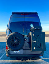 Rear of a gray RV rental van with a spare tire and storage box. Parked at a sunny beach, ready for adventure.