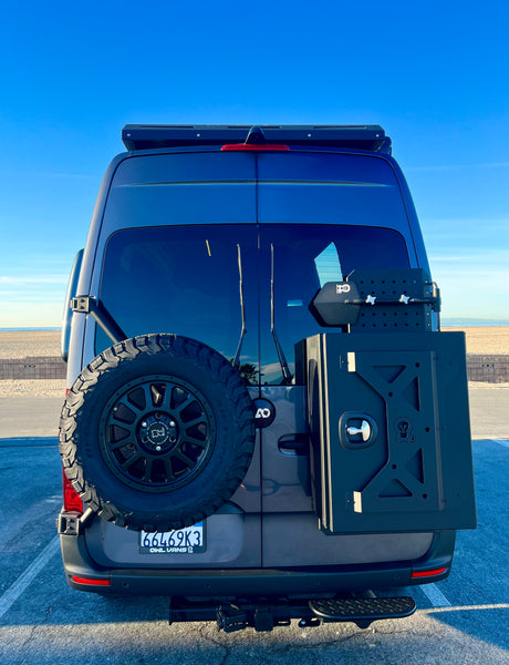 Rear of a gray RV rental van with a spare tire and storage box. Parked at a sunny beach, ready for adventure.