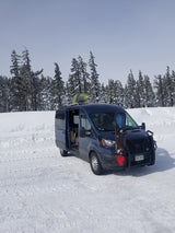 Blue van with snowboard & kayak, ideal for winter rv rental adventures in snowy forests.