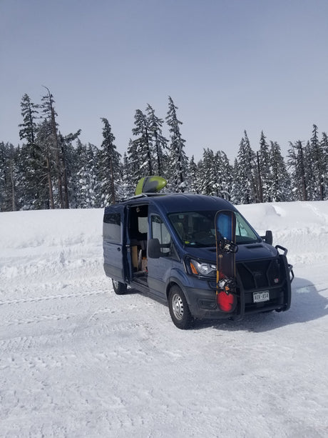Blue van with snowboard & kayak, ideal for winter rv rental adventures in snowy forests.