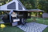 This grey and black rv rental pop-up camper is set up outdoors with an extended awning, outdoor kitchen, chairs, and mat.